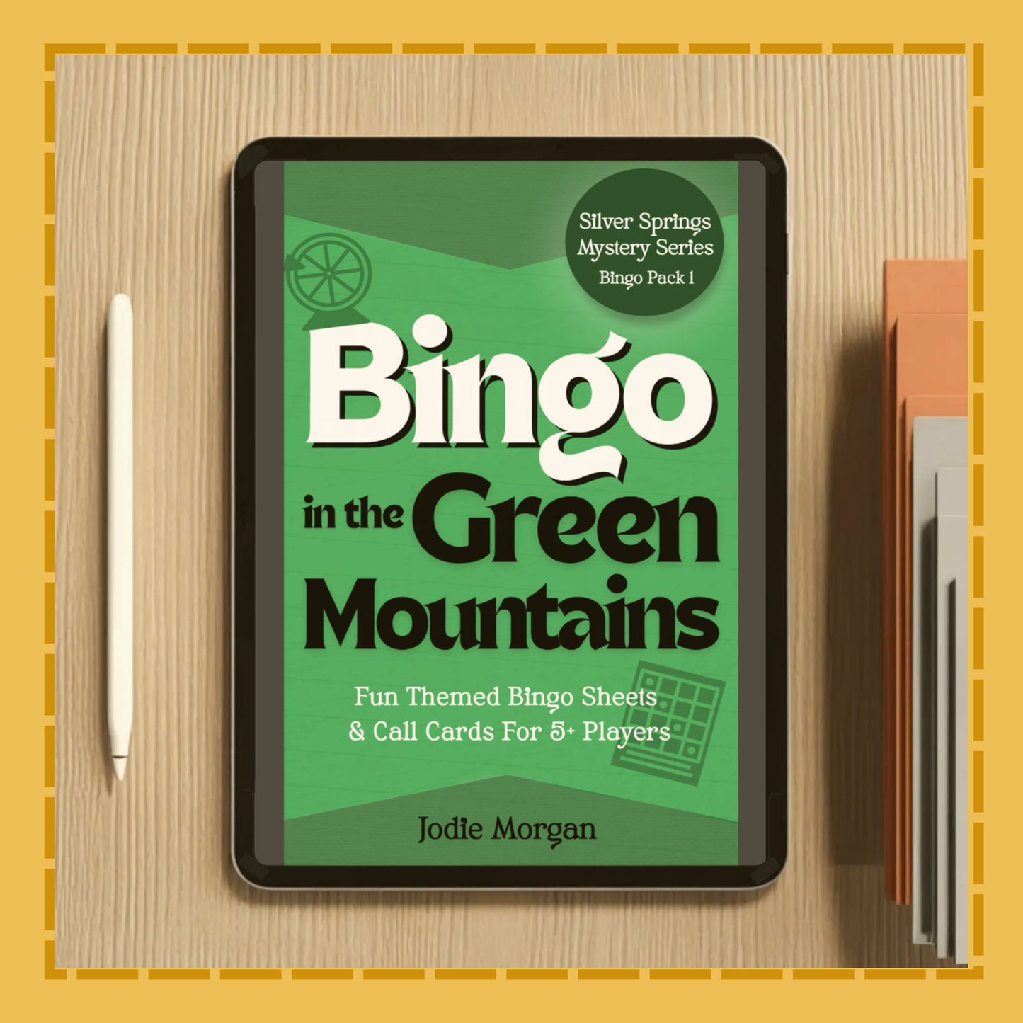 Digital book cover of 'Bingo in the Green Mountains' on a tablet with a pen and notebook on a wooden surface.
