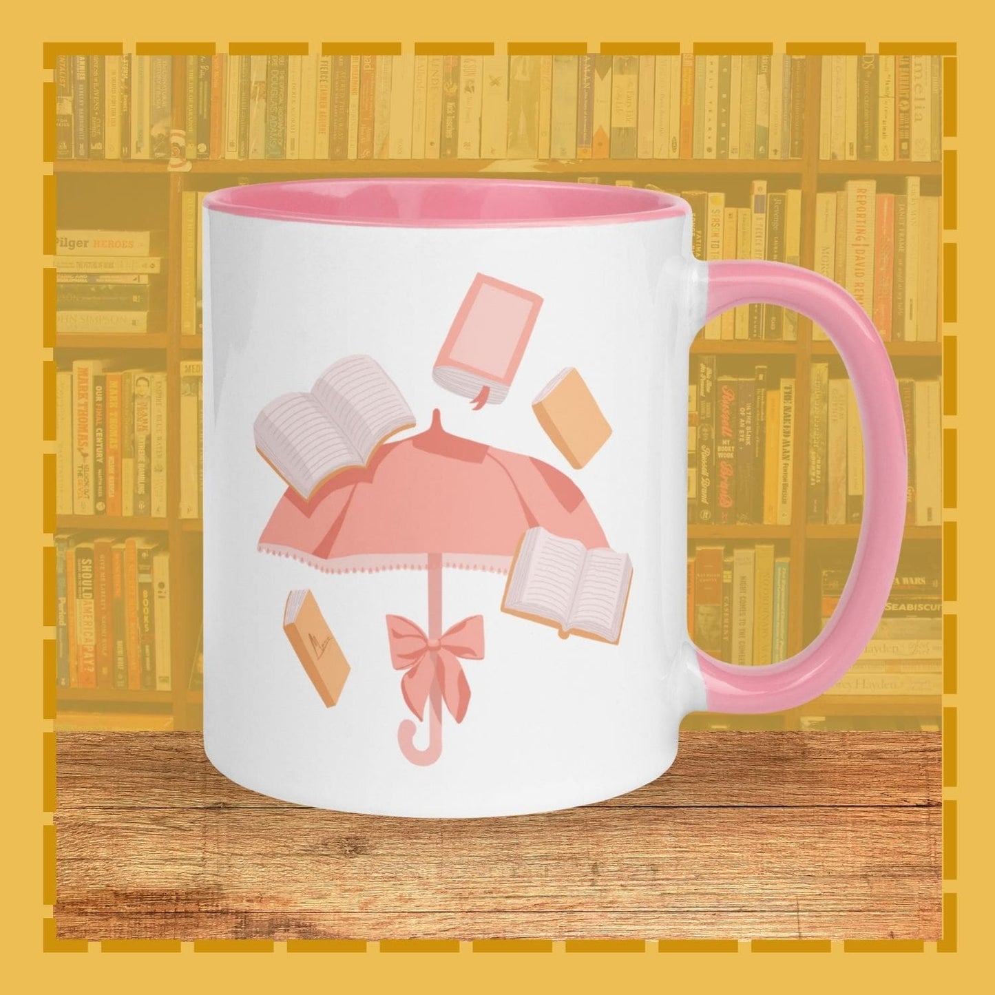 White mug with pink handle and interior, featuring a design of an umbrella and books, on a wooden surface with a bookshelf background.